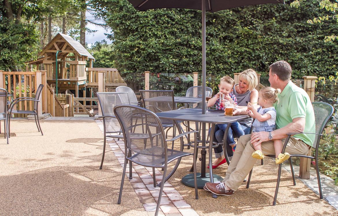 Family sat on the patio area enjoying a drink in the sunshine with the playground in the background