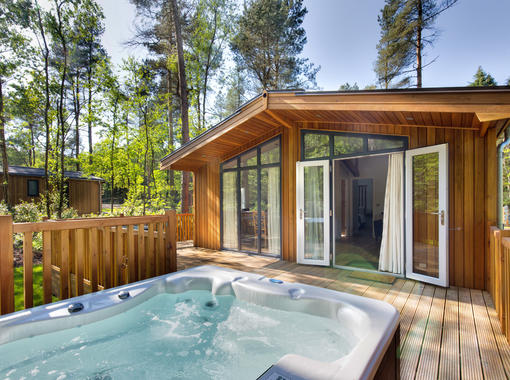 Lodge set within pine trees with beautiful blue sky, doors from lodge open onto verandah with out door hot tub