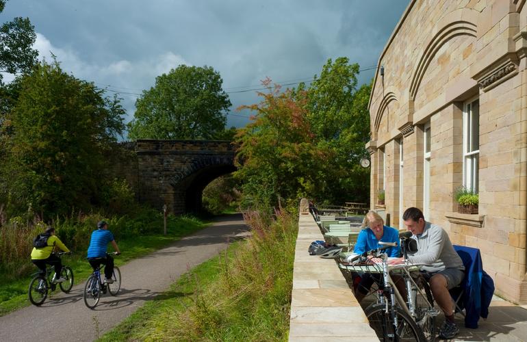 People cycling along the monsal trail at the side of the Hassop station cafe with people sat outside enjoying a bite to eat
